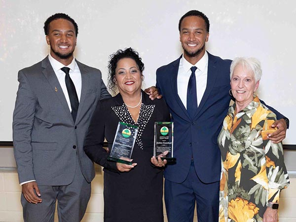 photo of two tall black students in suits with awards and two women in business attire