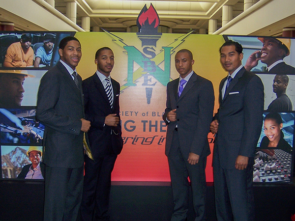 photo of four young men in suits standing in front of a NSBE banner