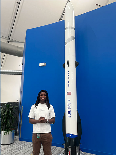 man with braids smiling in white tee and brown pants standing next to blue origin rocket