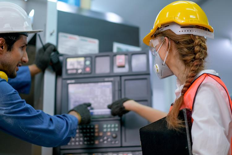photo of two people in hard hats looking at a control panel in a lab, from behind