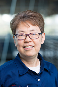 A woman with short dark hair and glasses looks at the camera with a slight smile, wearing a dark blue collared shirt, with a softly blurred indoor background.