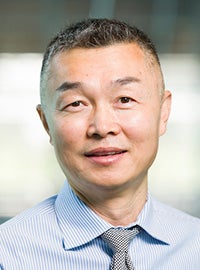 man with short dark hair wearing blue striped collared shirt and gray checked tie smiles at camera