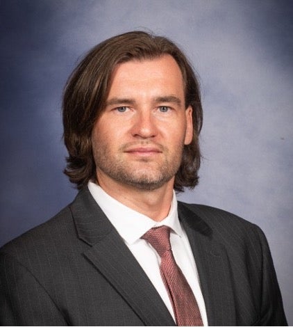 man with chin-length brown hair white collared shirt gray jacket and rose colored tied looking at camera against blue background