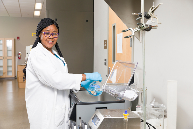 woman with long black braids wearing white lab coat and blue medical gloves works in clean gray lab pouring clear liquid from a bulb beaker into another container over a scientific instrument with clear lid while she looks at camera while wearing glasses and smiling