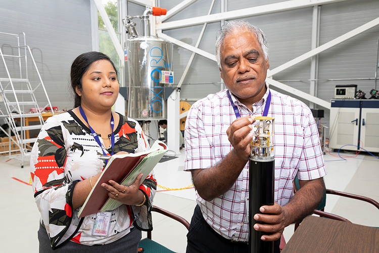 Two researchers smile while examining a small component on a cylindrical lab instrument in a research facility.