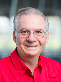 Portrait of a smiling man with glasses and gray hair wearing a red collared shirt against a softly blurred indoor background