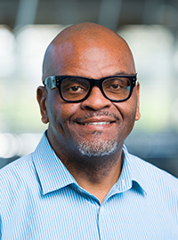 Portrait of a man wearing black-framed glasses and a light blue striped button-down shirt, smiling, against a softly blurred indoor background.