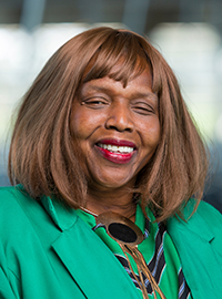 Portrait of a smiling woman wearing a green blazer and gold necklace, standing indoors against a softly blurred background