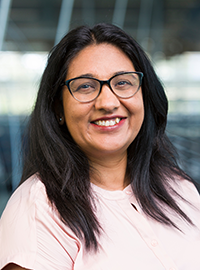 Portrait of a smiling woman with glasses and long dark hair wearing a light pink blouse against a softly blurred indoor background