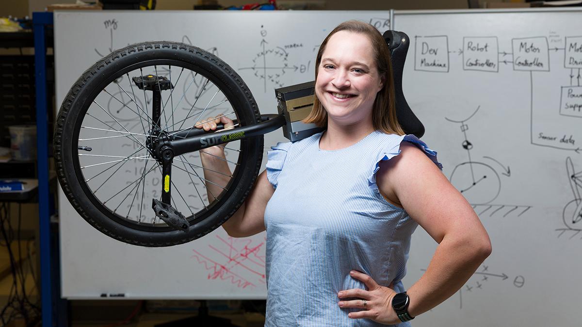 photo of taylor higgins phd with unicycle over her shoulder in front of math-filled white board at famu-fsu engineering