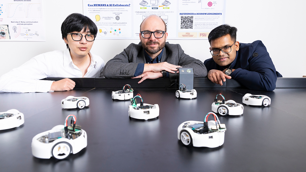 Three researchers lean over a table surrounded by multiple small robots during a laboratory demonstration with research posters in the background.