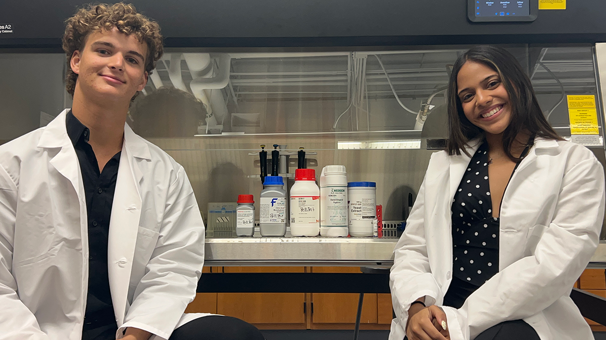 Two students in white lab coats sit on either side of a biosafety cabinet in a lab. Inside the cabinet, pipettes hang above lab supply containers, including bottles hand-labeled “PelliTect.”