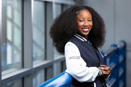 A young woman with curly hair smiles while leaning against a blue railing, wearing a navy and white jacket with Boeing TMFC Scholar Program text on the sleeve.