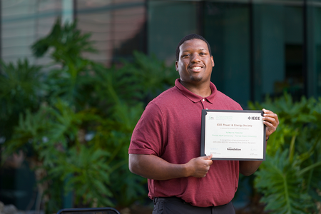 A student stands outdoors holding an IEEE Power & Energy Society scholarship certificate while smiling toward the camera.