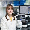 A woman sitting in a lab smiles at the camera in front of a microscope with a screen.