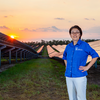 photo of woman in blue shirt and white pants standing in front of solar panel array at sunset