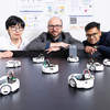 Three researchers lean over a table surrounded by multiple small robots during a laboratory demonstration with research posters in the background.