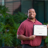A student stands outdoors holding an IEEE Power & Energy Society scholarship certificate while smiling toward the camera.