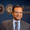 photo of man in glasses blue shirt gray tie smiling against dark background