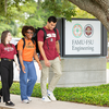 Three students walk together on a campus sidewalk beside a sign reading “FAMU-FSU Engineering.” The students wear university apparel and backpacks while talking beneath a large oak tree.