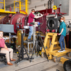 Three students pose around a large red experimental apparatus in an engineering laboratory. One sits at left, one stands on a platform beside the equipment, and one stands on a yellow rolling platform at right.
