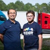 Two men stand outdoors in front of semi-trucks parked side by side, with trees and a clear sky in the background. The man on the right wears a navy RIDER T-shirt.