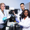 Three researchers wearing white lab coats and protective eyewear pose around a laboratory microscope. A woman on the left holds a notebook and pen, a man behind the microscope adjusts the equipment, and a woman on the right smiles while holding a clear sample tube.