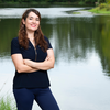 Woman in a navy blouse standing with arms crossed in front of a calm lake, smiling at the camera.